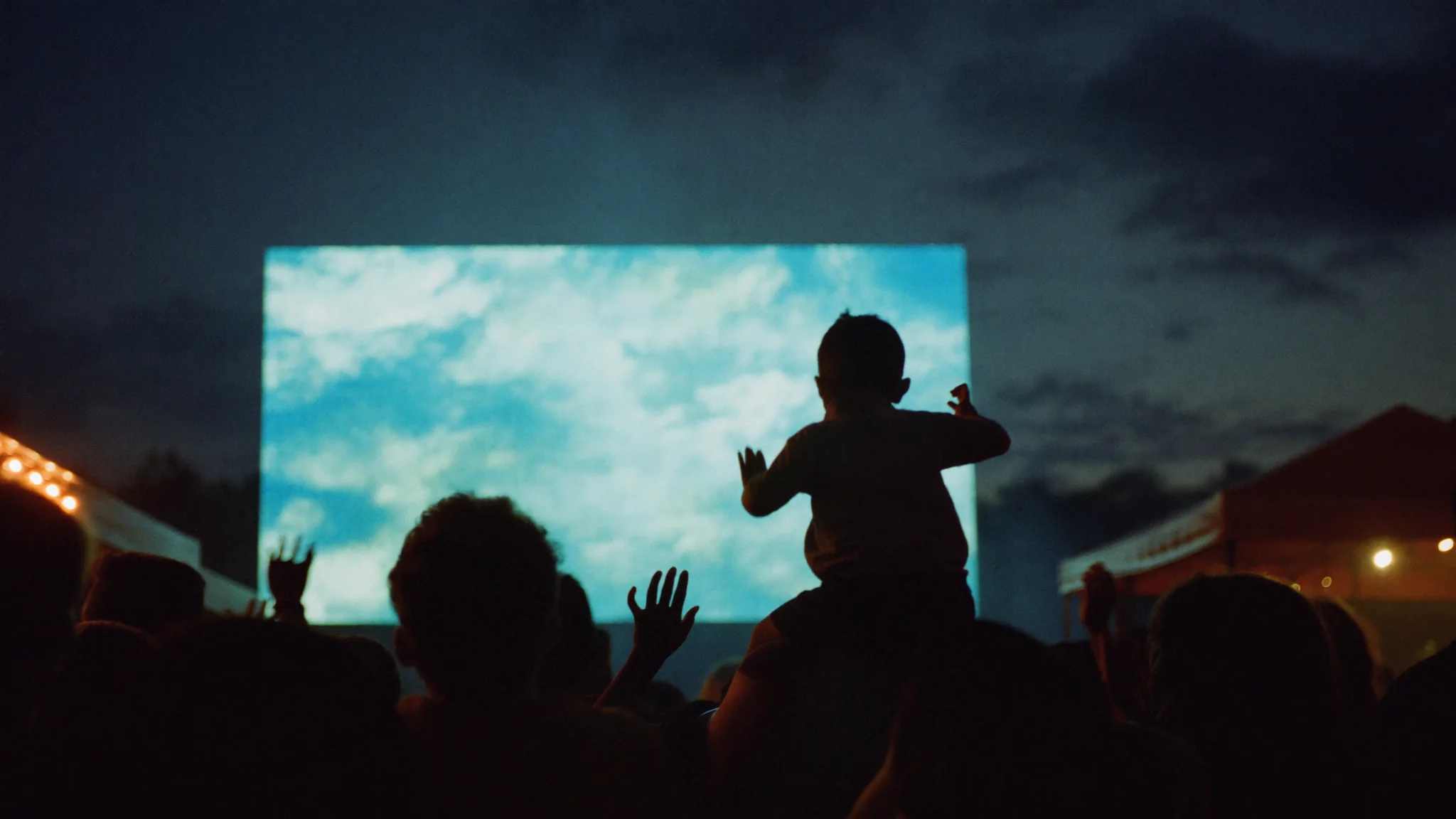 An outdoor watch-party market at dusk, a giant screen behind a curated local market.